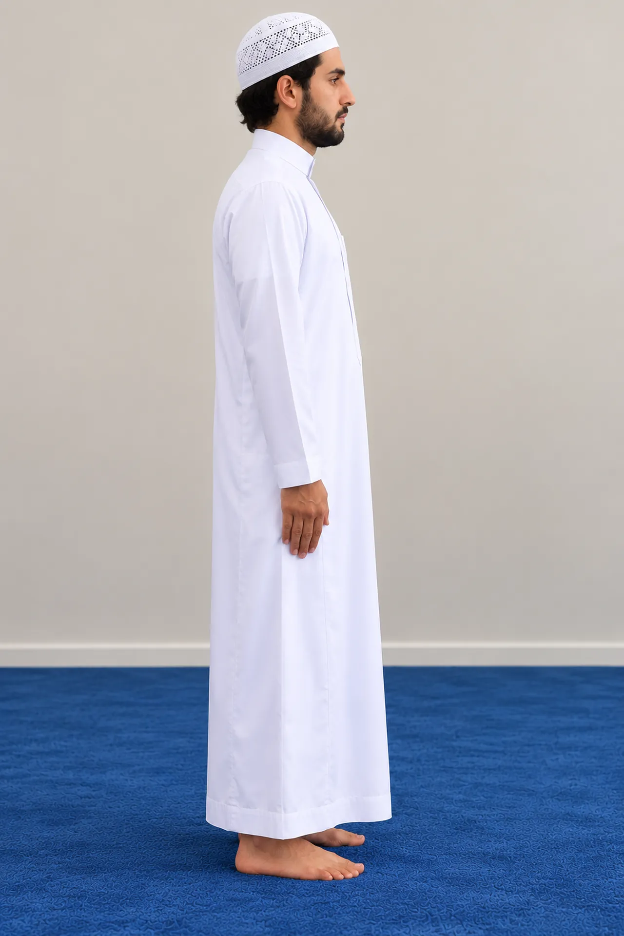 Le même homme en qamis blanc et kufi blanc, debout de profil sur le tapis bleu et orienté vers la droite, dans une posture calme, le regard baissé.